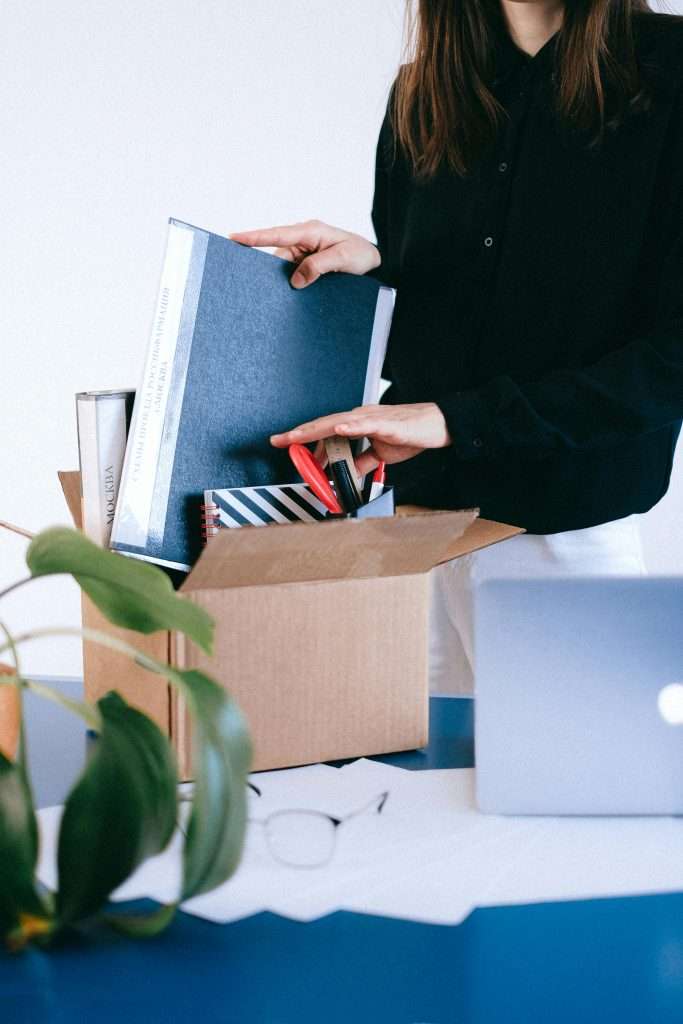 Adult woman organizing office supplies into a cardboard box, possibly symbolizing job change or relocation.